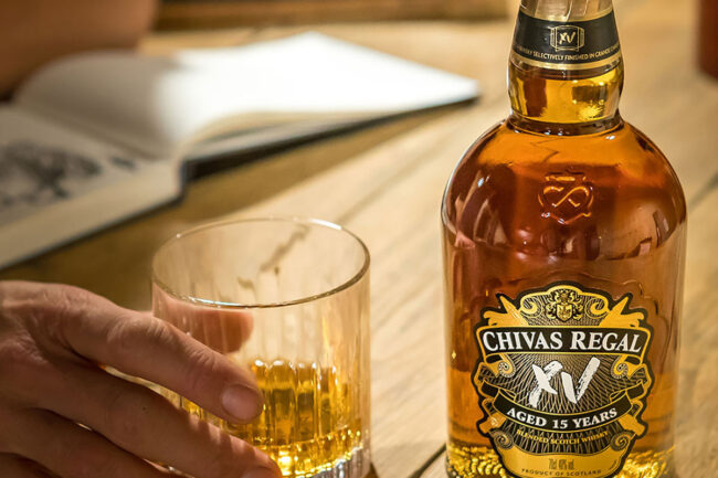 Hand holding glass of whisky beside Chivas Regal 15 Years bottle on table.